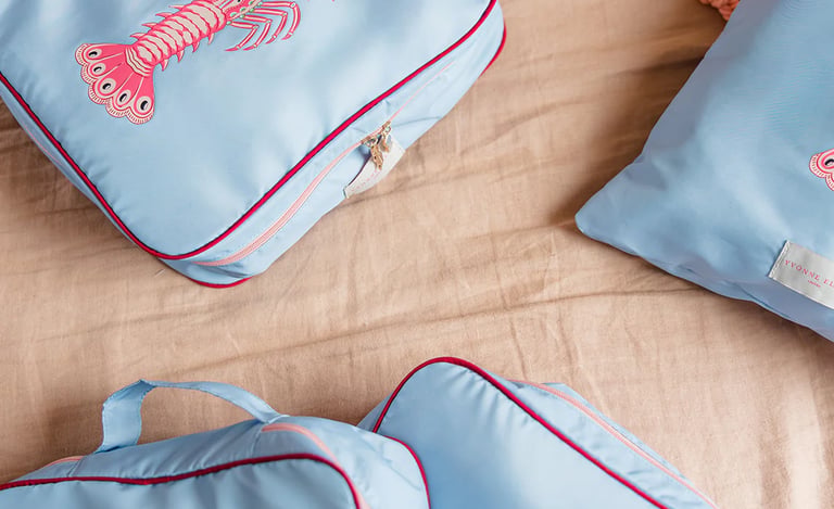 Light blue travel packing cubes with pink lobster embroidery on a beige linen surface.