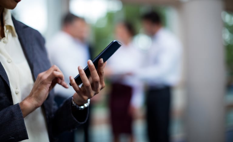 Person using a smartphone with colleagues conversing in the background.