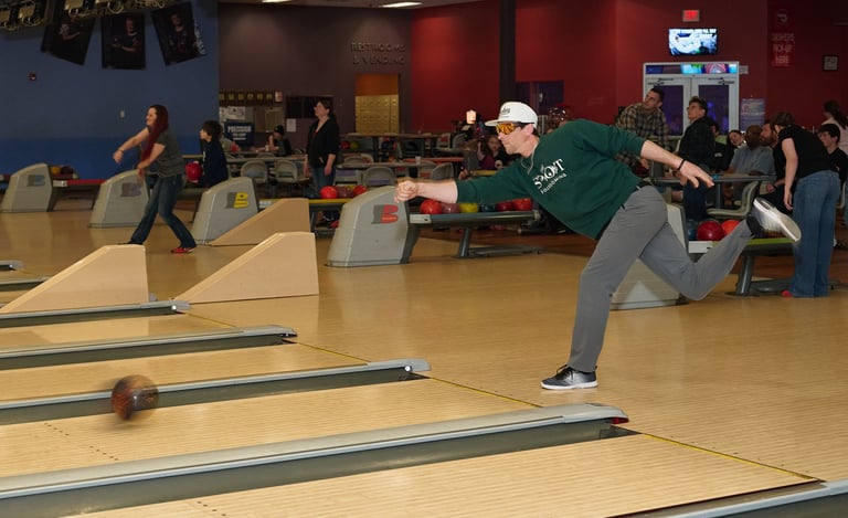 A male bowler in a green shirt releases a bowling ball down a wooden lane at a bowling alley.