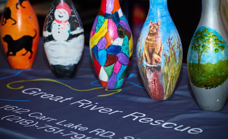 Hand-painted bowling pins featuring art of a cat, snowman, and nature on a Great River Rescue table.