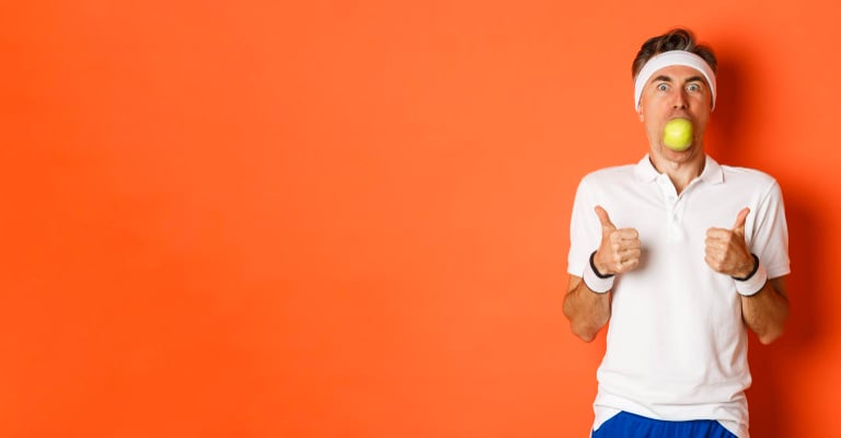 Middle-aged man in workout gear holding a green apple in his mouth giving a thumbs up on orange background.