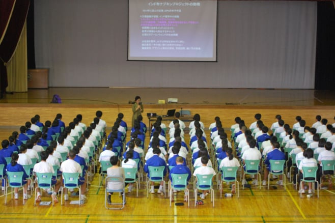 A school hall full of students with stage and large screen on stage.