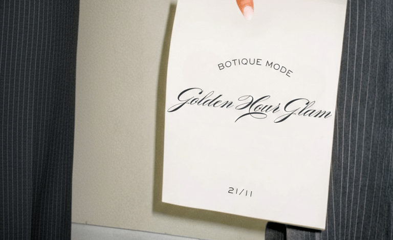 Mano sosteniendo una invitación Golden Hour Glam de Botique Mode sobre un pantalón de raya.