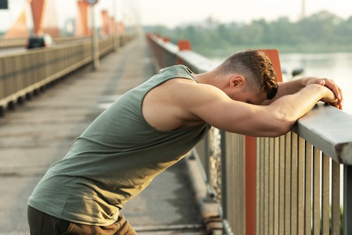 Athletic man in tank top leaning on a bridge railing while resting after an outdoor workout.