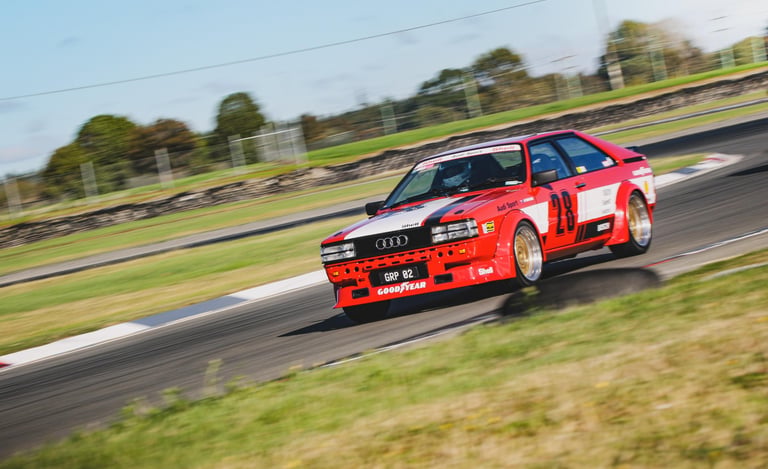 Red Audi Quattro race car with Goodyear branding speeding on a paved motorsport track.