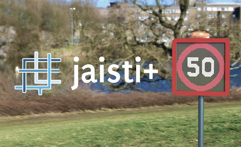 Jaisti+ logo next to a 50 speed limit sign with a blurred park and lake background.