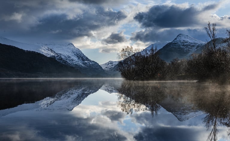 Snowy mountains reflecting in a calm, misty lake under a dramatic cloudy sky.