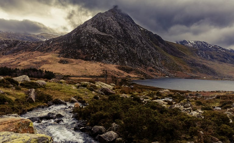 Dramatic mountain peak overlooking a scenic lake and rocky stream in Snowdonia, Wales.