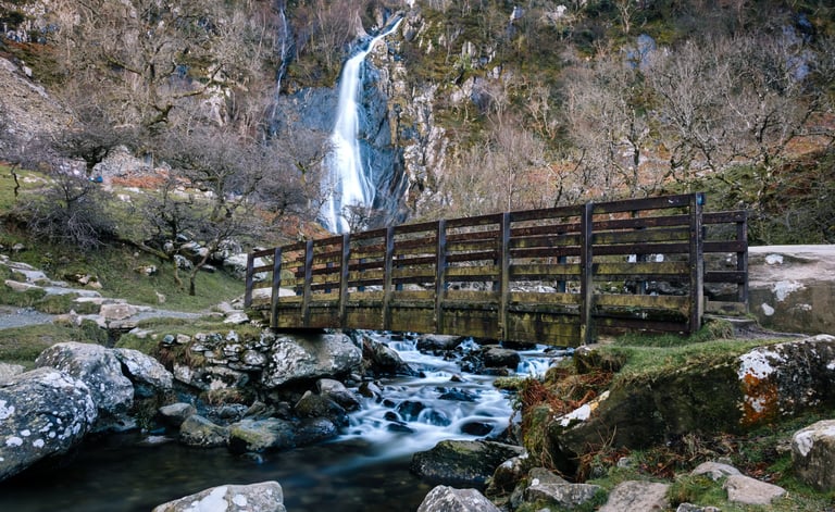 A wooden footbridge crossing a rocky stream with the Aber Falls waterfall in the background.