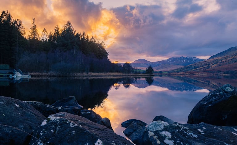 Scenic sunset reflection over a calm mountain lake with pine forest and rocky shoreline.