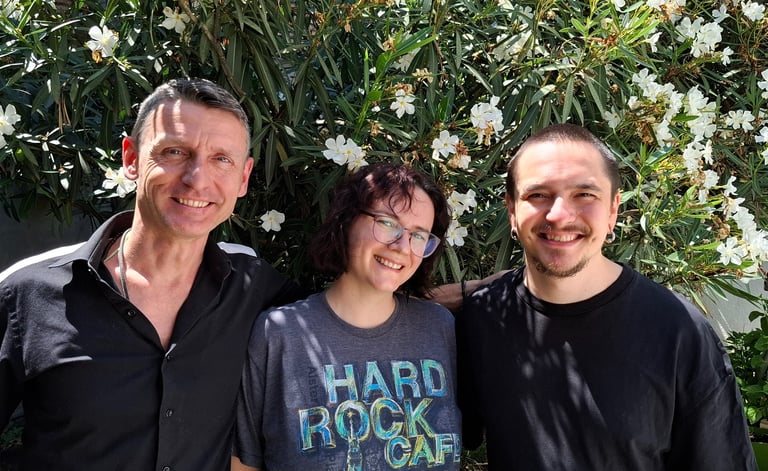 Three smiling friends posing together outdoors in front of a blooming white oleander bush.