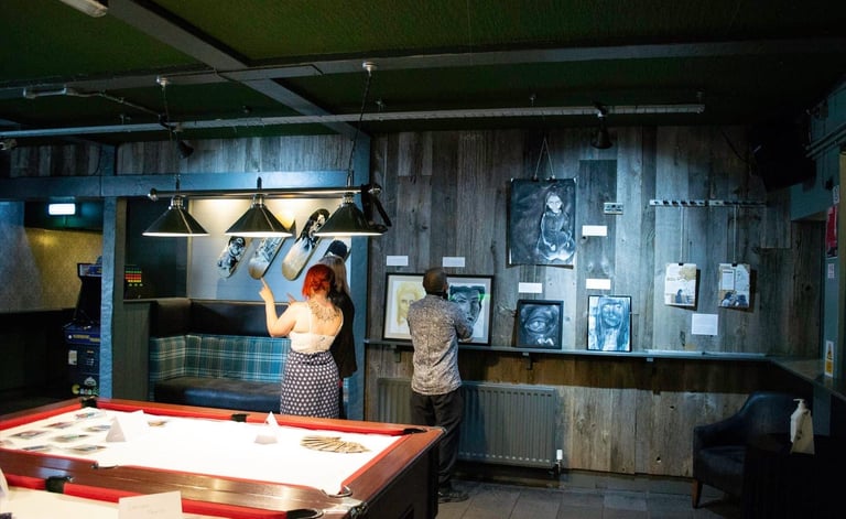 People at the exhibition looking and talking about art. Low lighting. Pool table in centre of room.