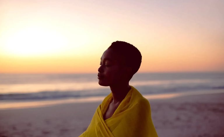 Une femme en foulard jaune se tient sur la plage au coucher du soleil, profitant du calme et de la beauté du moment.