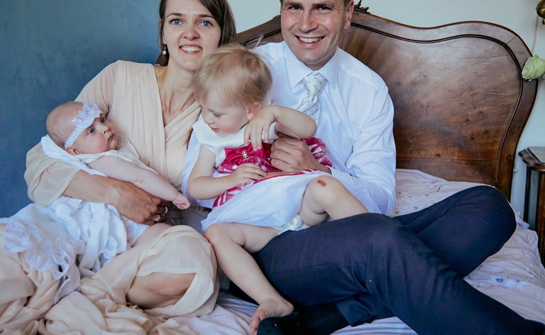 Kobieta i mężczyzna leżą na łóżku wraz z dziećmi. Przygotowania do Chrztu Świętego.