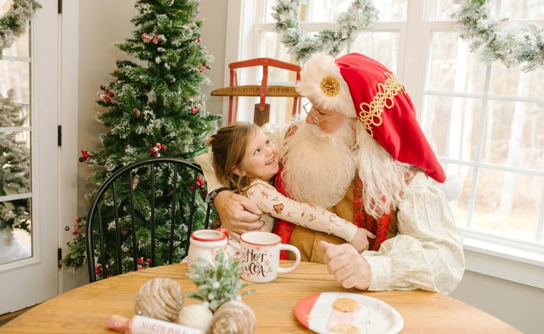 A smiling young girl hugging Santa Claus at a holiday table with cookies, cocoa, and a Christmas tree.