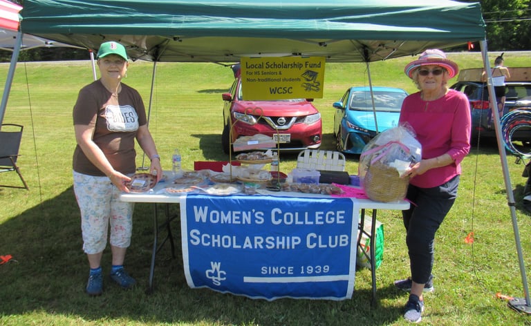 photograph of two women standing at a bake sale table for College Scholarship Club under small tent