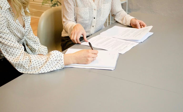 Two business women reviewing paper documents and taking notes during a professional office meeting.