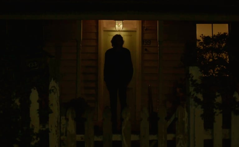 Dark silhouette of a person standing at a house door, under a porch light at night.