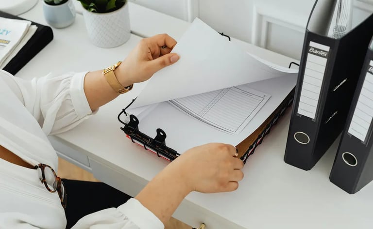 A person organizing office documents and paperwork in a wire tray on a white desk with binders.