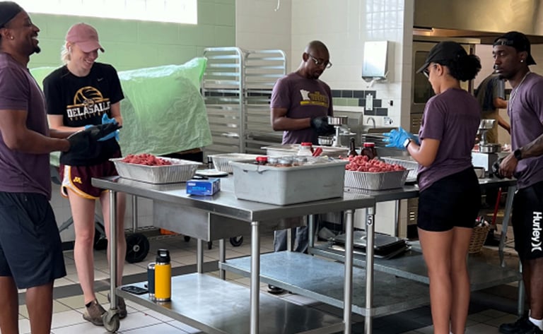 Volunteers in purple shirts preparing food together in a professional commercial kitchen setting.