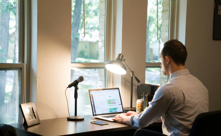 A man working at a home office desk with a laptop, microphone, and lamp by a window.
