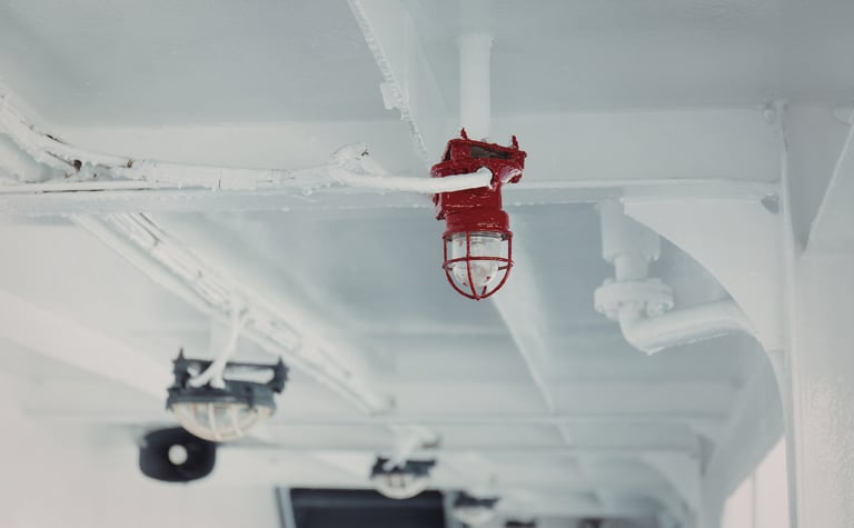 Industrial red marine cage light mounted on a white ship deck ceiling.