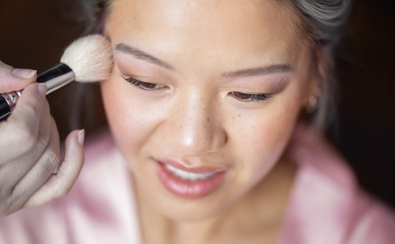 Une mariée en robe de soie rose recevant une application de maquillage de mariage avec un pinceau.