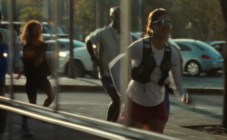 Athletic woman wearing a hydration vest and sunglasses running on a city street with other runners.