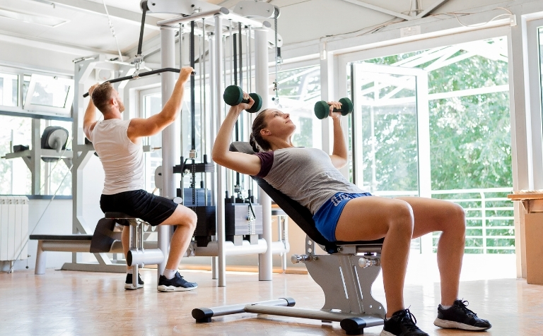 A man and woman perform strength training exercises using dumbbells and a lat pulldown machine in a bright gym.