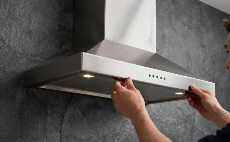 A close-up of a sleek designer kitchen chimney being mounted on a dark slate grey wall. Professional hands are visible ensuring precise alignment. Premium lighting highlighting the brushed metal finish.