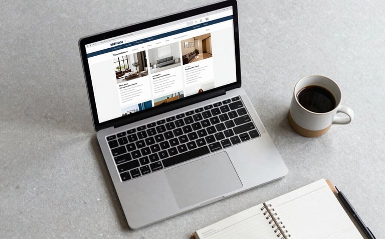 A top-down view of a designer's workspace featuring a modern laptop showing a clean website dashboard, a ceramic cup of coffee, and a minimal planner on a pale pebble gray desk.