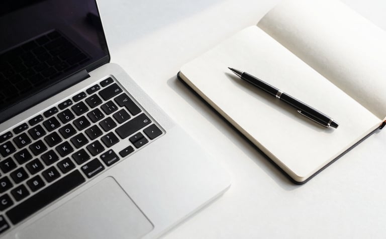 A top-down view of a modern minimalist desk featuring a sleek silver laptop, a white notebook, and a single pen. The lighting is bright and airy with soft shadows, incorporating steel blue and soft off-white tones to reflect a clean web design workspace.