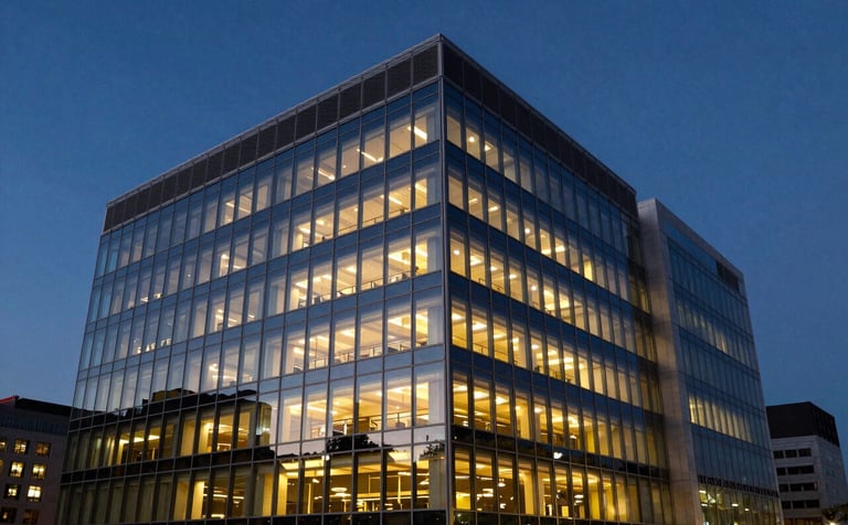 A wide-angle professional photograph of a modern glass-fronted North American / US commercial office building. The architecture is sleek, reflecting a dark navy blue sky at twilight with warm gold interior lighting glowing through the windows.