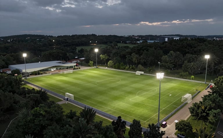 An aerial cinematic view of a modern football academy complex in Portugal at dusk, the floodlights illuminating the lush green pitches against a sophisticated deep forest green and slate grey sky.