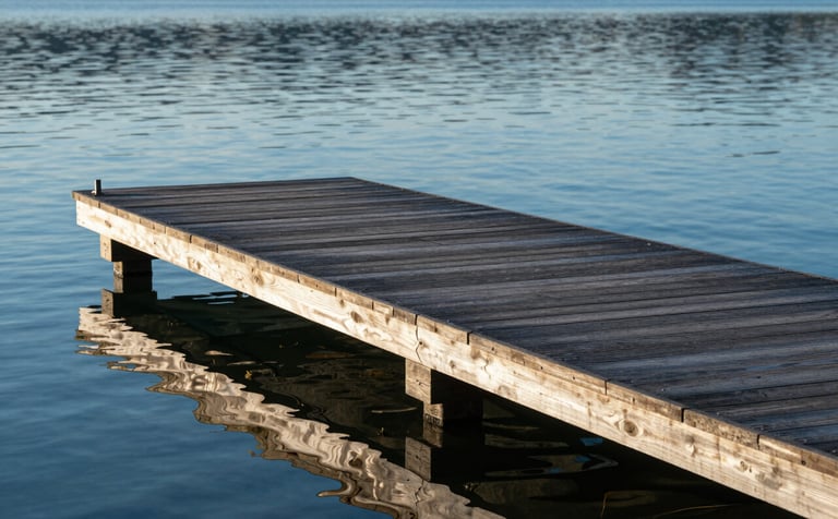 A high-end custom wooden dock with precision joinery extending into a calm blue lake. The timber has a professional, dark protective finish. Morning sun creates soft reflections on the water. The scene incorporates brand colors #1B3A4B and #4F7F8F in the shadows and wood tones.