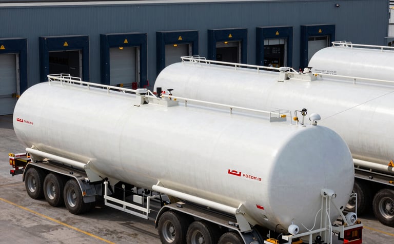 Clean, industrial photography of a fleet of white fuel tankers at a modern loading terminal. The environment is organized and highly professional, reflecting strategic supply chain efficiency in a Global / Professional setting. Colors emphasize dark navy and slate blue.