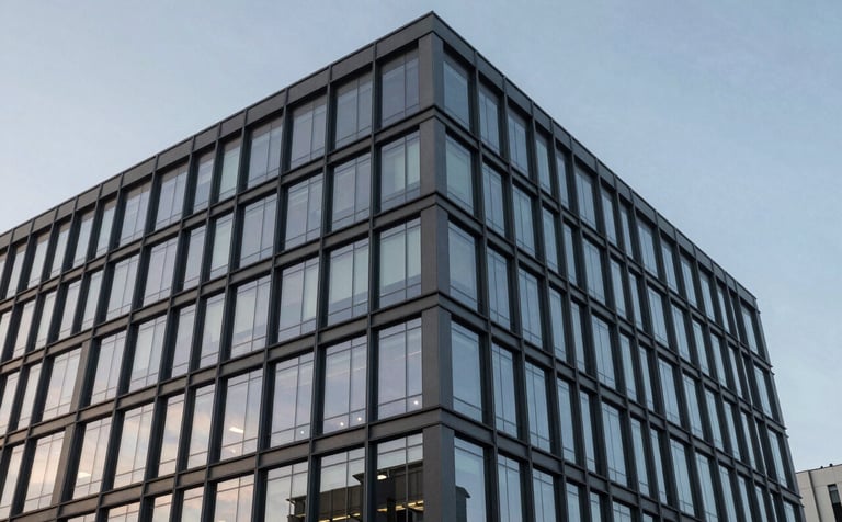 A crisp, eye-level photograph of a newly completed modern commercial office building with glass windows reflecting a soft blue-grey sky. The building features clean lines and a professional, sophisticated aesthetic.