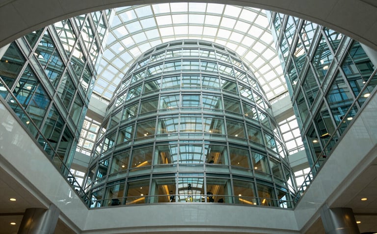 Low angle photography of a modern, glass-domed North American government building atrium. The scene is bright and airy, reflecting innovation and transparency with light blue and grey tones throughout the architecture.