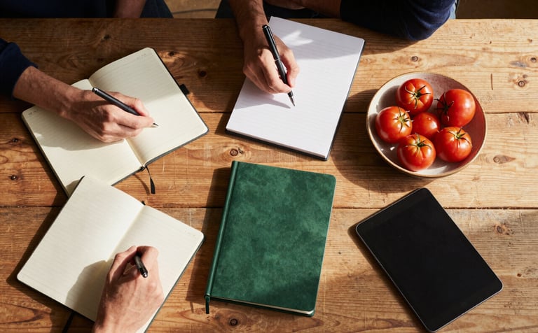 Top-down view of a rustic wooden table where a marketing team is planning a campaign. There are notebooks, a tablet, and a ceramic bowl of small red tomatoes. Warm natural sunlight filters in, highlighting Matte Forest Green and Deep Ripe Crimson accents. Scandinavian minimal style.