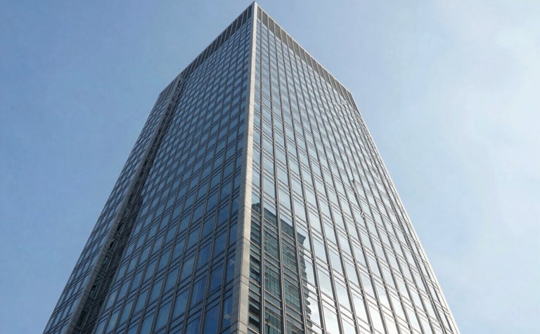 A wide-angle, low-angle photograph of a modern glass skyscraper reflecting a clear blue sky. The architecture is sharp and modern, representing global financial hubs. The colors are predominantly Soft Ice White and Slate Blue, conveying an authoritative and global perspective on the economy.