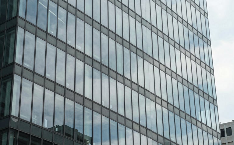 A sophisticated architectural photograph of a modern glass office building in a North American city. The glass reflects a soft, cloudy sky, creating a sense of digital flux and modern transformation. The colors are dominated by muted blue and light gray.