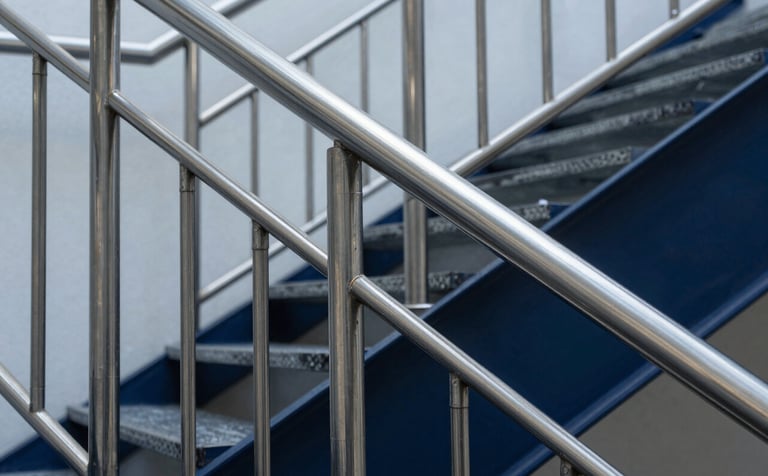 Close-up architectural shot of a precision-engineered stainless steel railing and staircase in a contemporary South American / Brazilian commercial building. The steel blue and dark navy reflections of the metal are sharp. The lighting is studio-quality, emphasizing the brushed texture and professional finish of the inox projects.