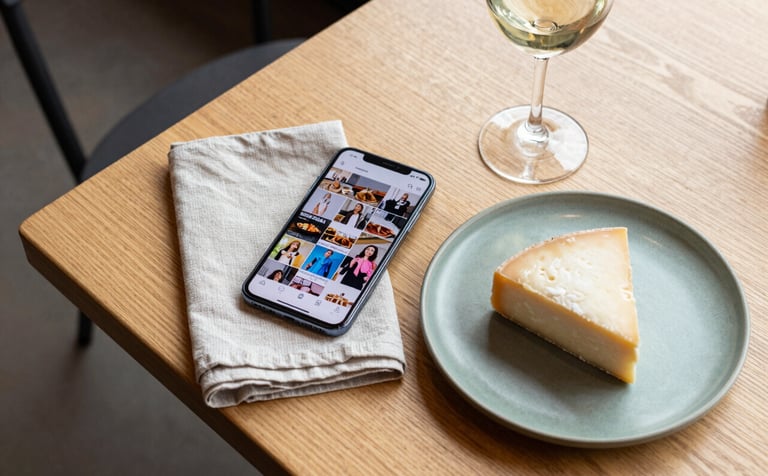 A flat lay photography shot on a light wood Scandinavian-style table in a North American cafe. A smartphone showing a vibrant social media feed sits next to a linen napkin, a matte green ceramic plate with artisanal cheese, and a glass of wine. Sophisticated, bright natural lighting, professional and clean composition.