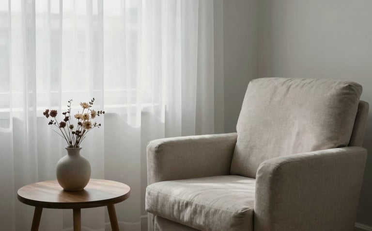 Photography of a peaceful therapy room corner. A soft-textured armchair sits by a window with misty white curtains. On a side table, there is a small ceramic vase with dried flowers. The lighting is soft and natural, featuring tones of misty white and soft sage.