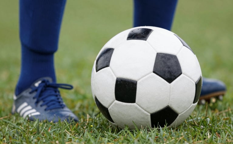 A close-up of a professional size 5 soccer ball on a lush green field in Quebec, with a player's feet nearby wearing socks in #4B657D blue. Clear, bright daylight, emphasizing community sports and quality equipment.