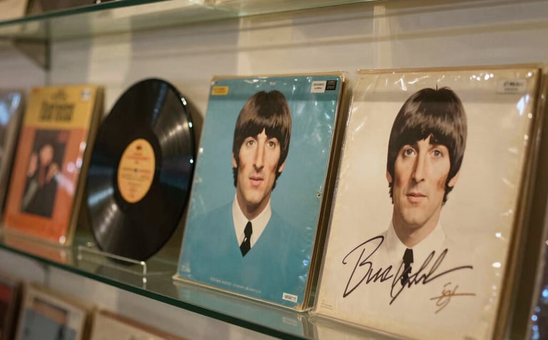 A close-up photograph of a rare Beatles record collection and signed memorabilia displayed behind polished glass in a South American gallery, with warm golden and muted teal tones.