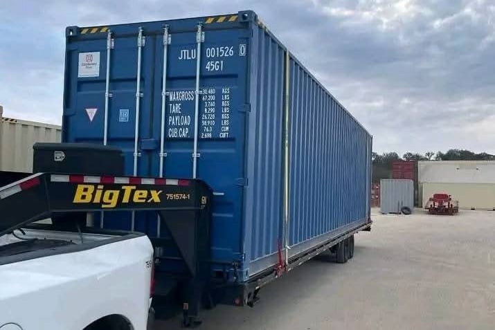 A blue shipping container loaded onto a Big Tex flatbed trailer attached to a pickup truck.