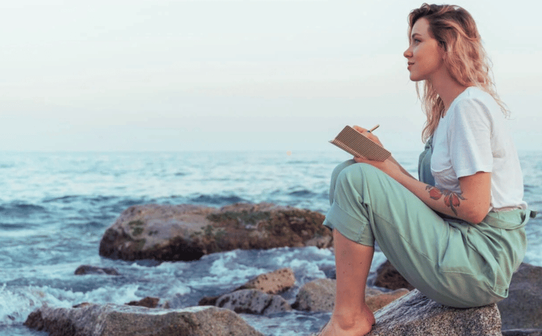 Woman by the ocean holding a journal and pen, reflecting on emotional intelligence & self-awareness.