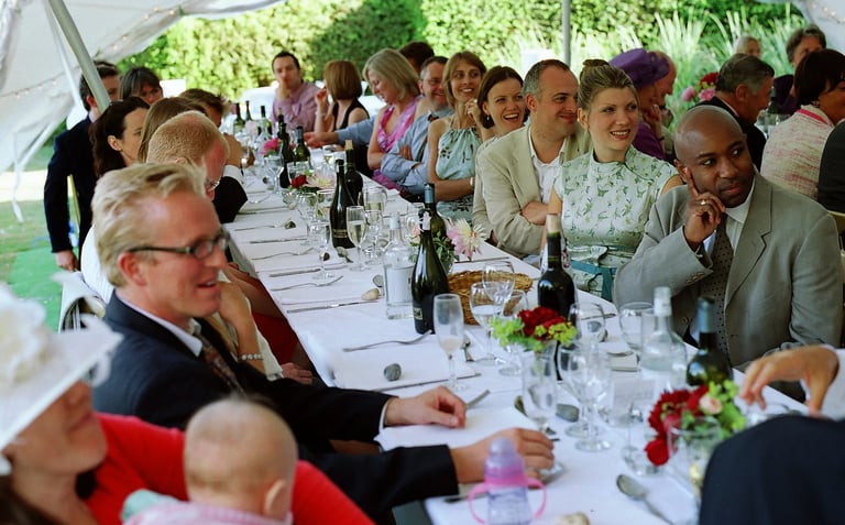 A diverse group of smiling wedding guests seated at long tables for an outdoor marquee reception dinner.