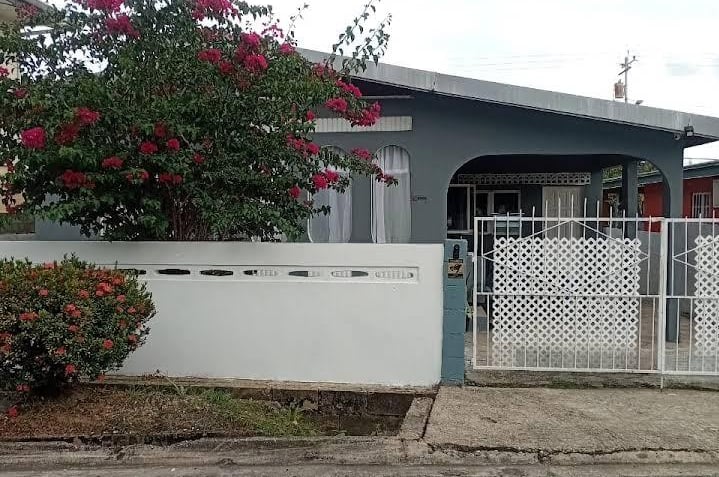 Small gray house with a white lattice gate and blooming pink bougainvillea tree out front.